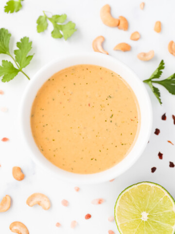A creamy orange salad dressing in a white bowl. There are supporting ingredients around the bowl including: cilantro, cashews, red pepper flakes, a sliced lime, and salt