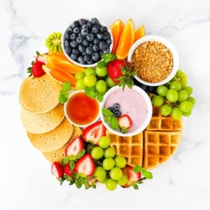 A round board filled with pancakes, waffles, fruit, yogurt, and syrup