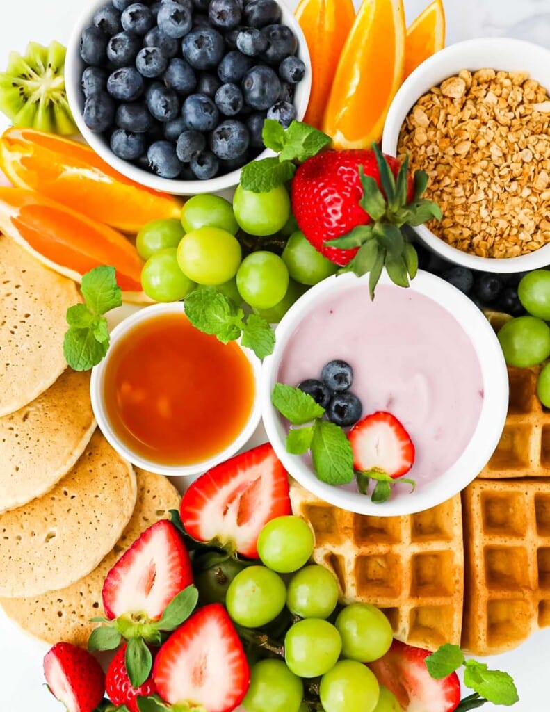 A close up picture of a brunch board filled with waffles, pancakes, yogurt, granola, and fruit
