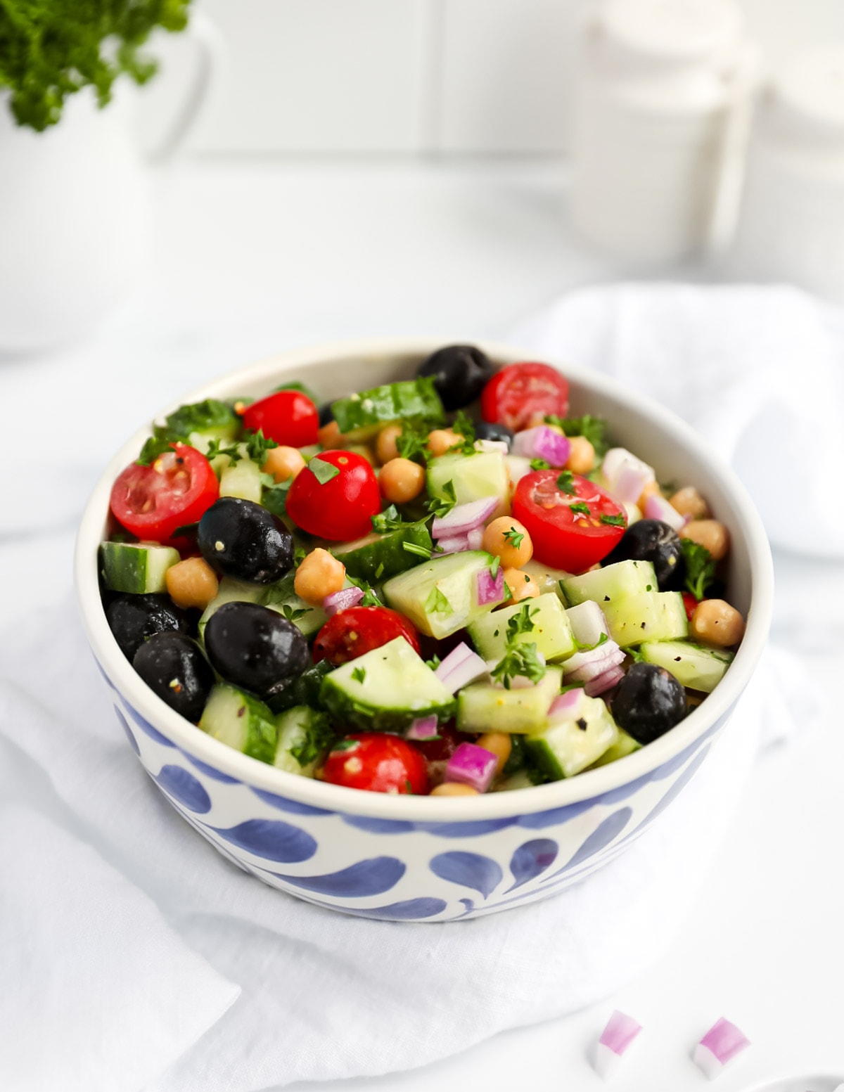 A blue and white bowl filled with a Greek salad including olives, tomatoes, chickpeas, cucumbers, and onion, covered in fresh herbs