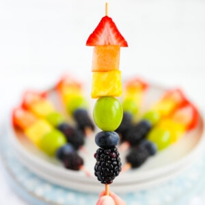 A hand holding up a wooden stick with one piece of strawberry, melon, pineapple, grape, blueberry, and blackberry