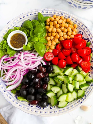A large salad with lettuce, basil, chickpeas, tomatoes, cucumbers, olives, red onion, and a small white dish filled with dressing