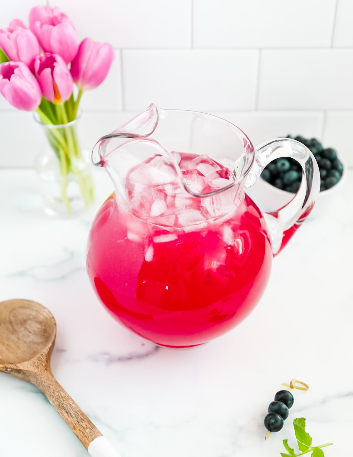 A clear glass pitcher filled with bright pink lemonade. There are ice cubes in the pitcher and flowers, berries, and a spoon outside