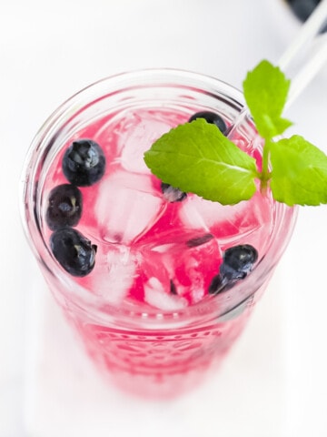 A clear cup filled with pink lemonade. There are ice cubes, berries, and a fresh sprig of bright green mint in the cup.
