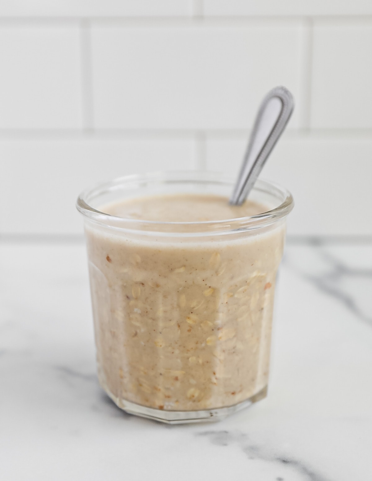 A clear glass jar filled with oats and oat milk with a spoon in it