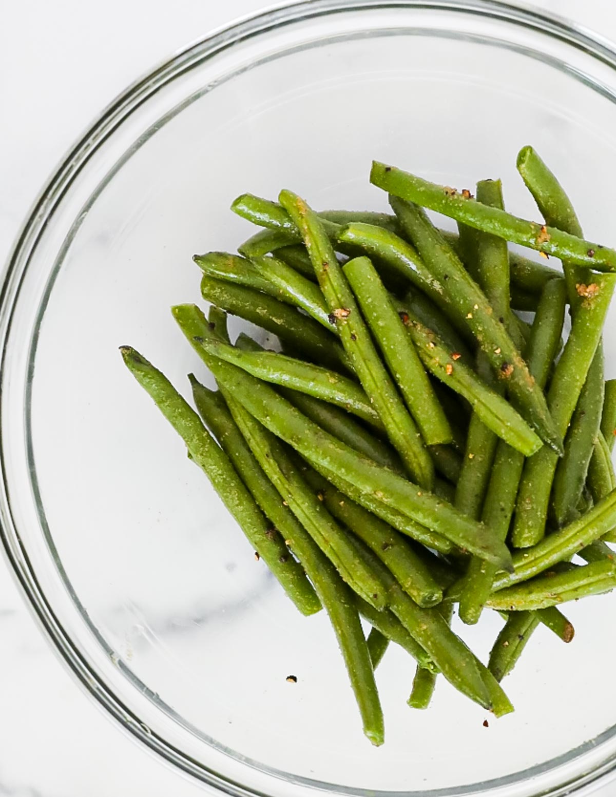 A clear glass bowl filled with fresh green beans covered in olive oil and seasonings.