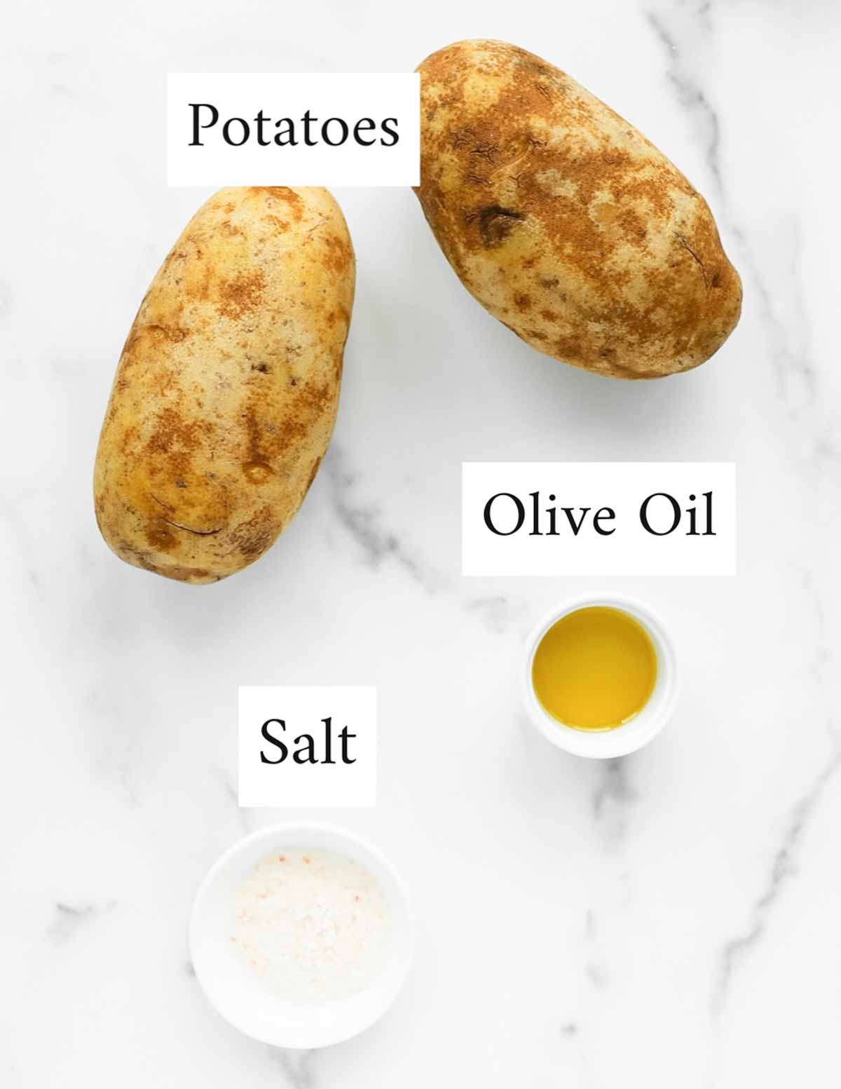 an overhead image of two potatoes, a small white dish containing olive oil, and a small white dish containing salt