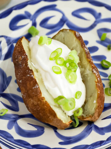 A crispy cooked potato on a blue and white plate. There is a large scoop of creamy sour cream and sliced green onions on the sliced potato.