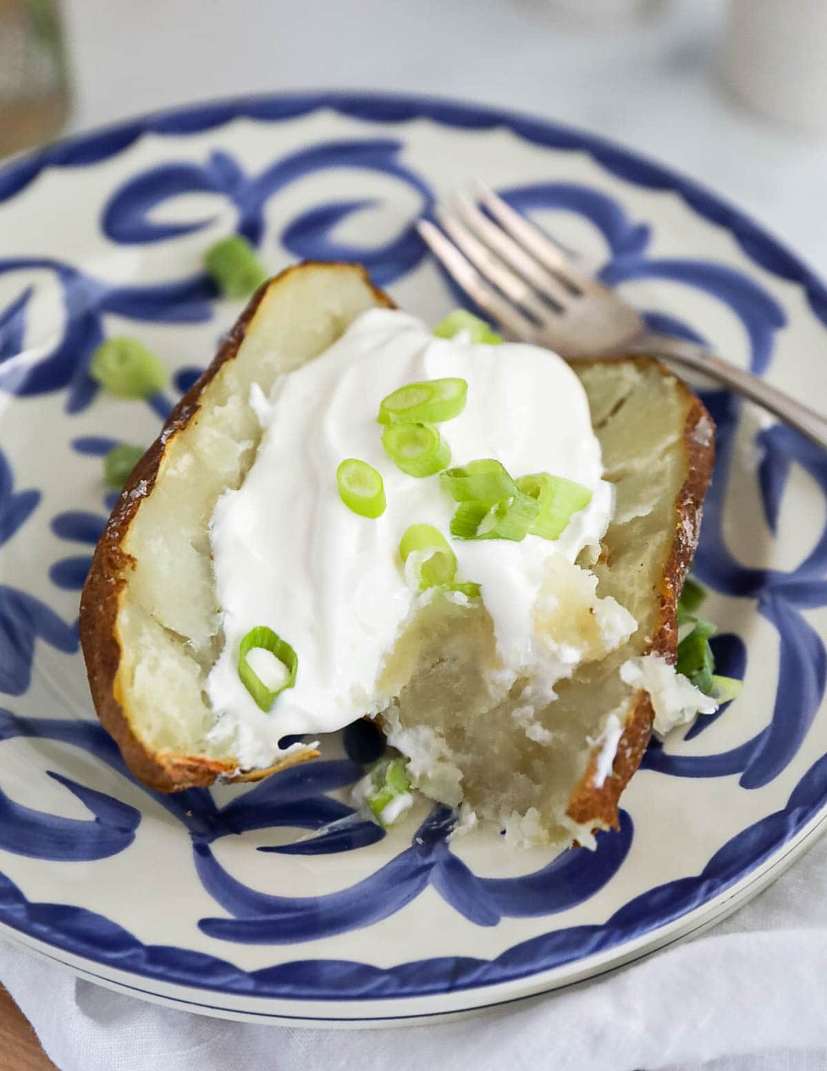 A baked potato on a blue and white plate. The potato is sliced in half, there is a dollop of sour cream and sliced scallions inside the potato, and a fork next to the potato.