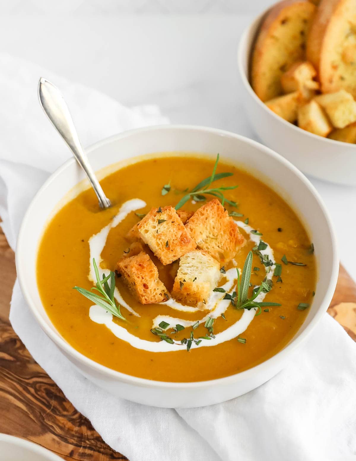 A large white bowl of orange squash soup with croutons, herbs, and coconut milk on top. There is a spoon sitting in the soup.