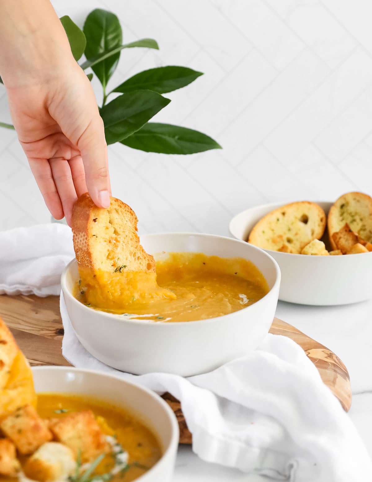 A hand dipping a piece of bread into a bowl of orange soup.