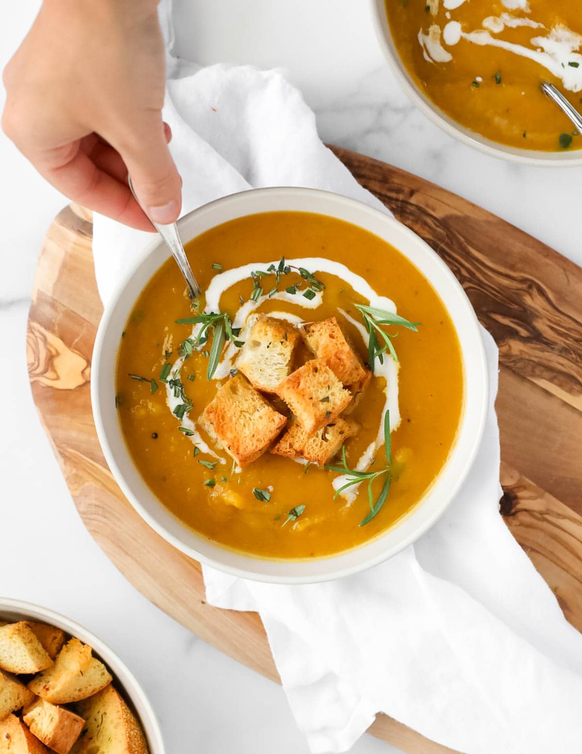 A hand dipping a spoon into a bowl of orange soup. There are herbs, croutons, and coconut milk on top of the soup.
