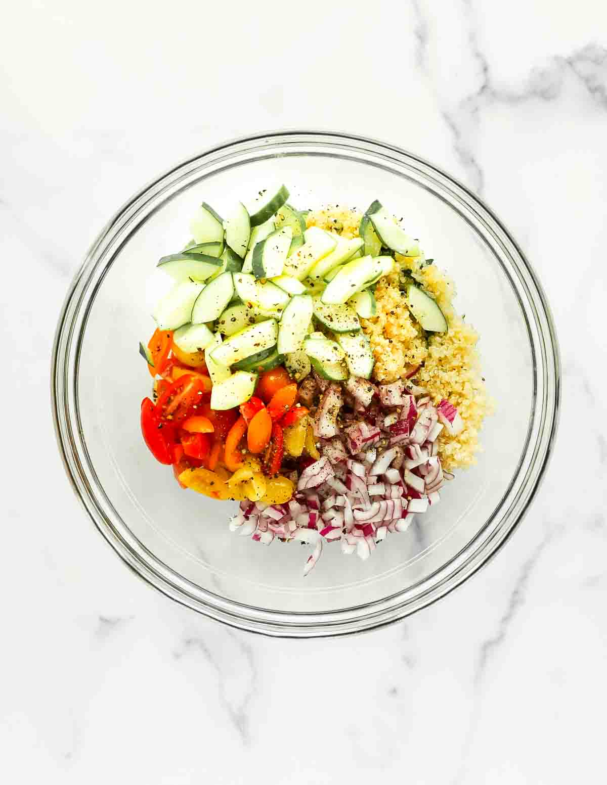 a clear mixing bowl filled with cucumbers, quinoa, red onion, tomatoes, and pepper on top