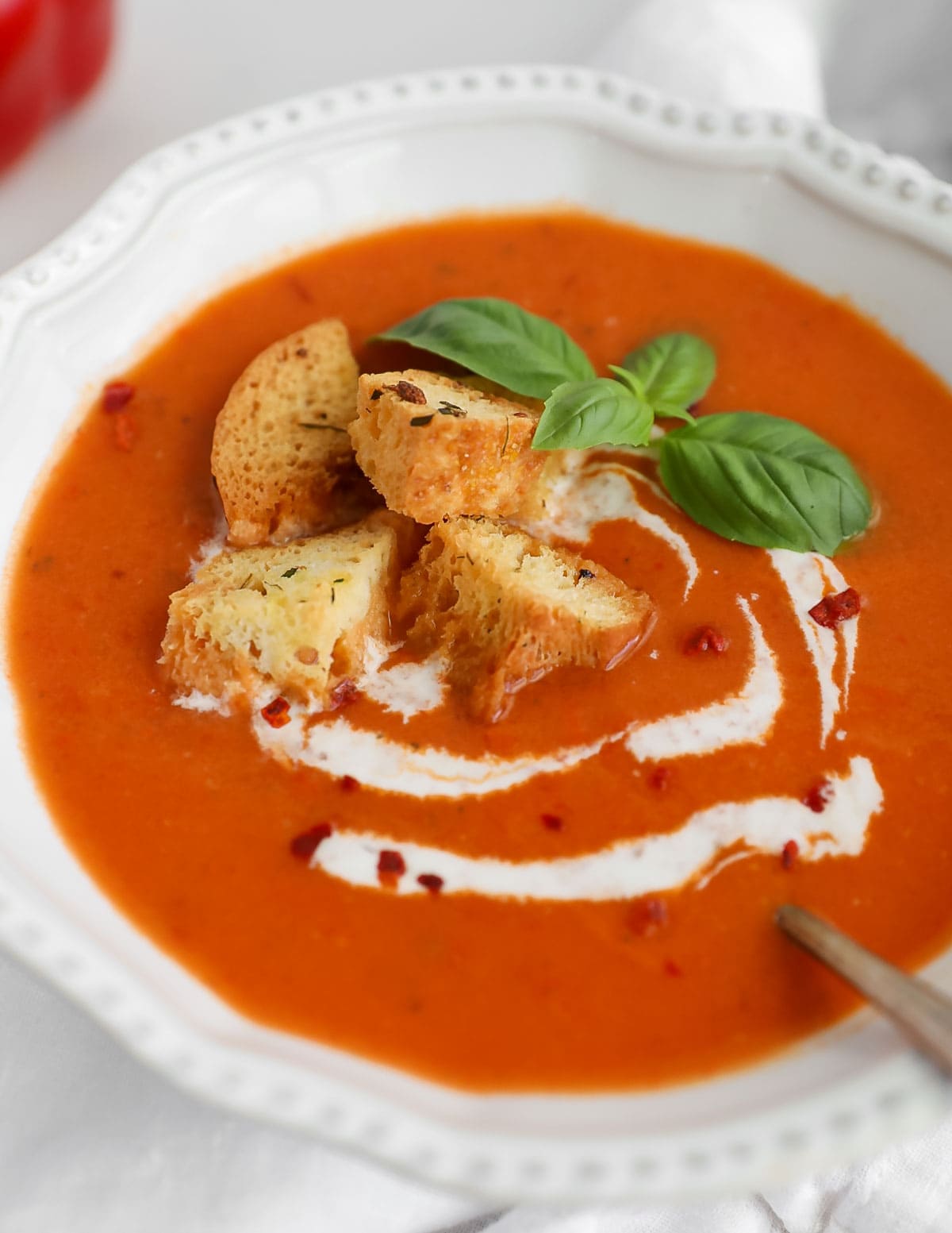 A close up image of creamy red soup in a white bowl. On top of the soup there are four croutons, basil leaves, white coconut milk, and crushed red pepper flakes.