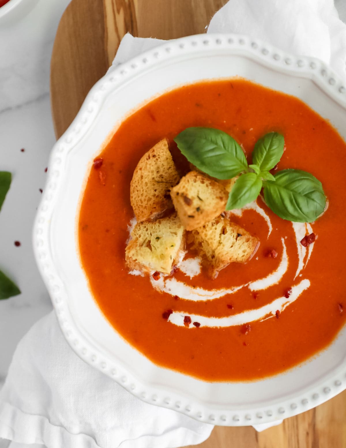 An overhead image of red soup in a white bowl garnished with croutons, basil, coconut milk, and crushed red pepper flakes.