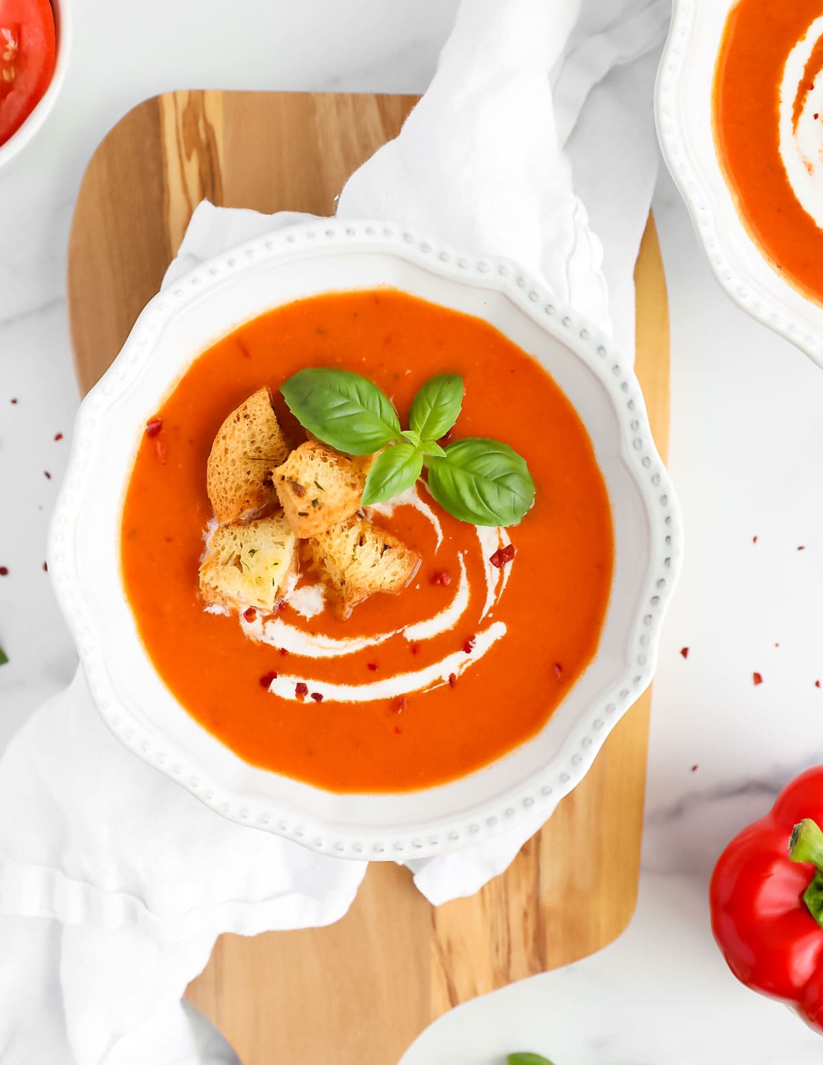 An overhead image of red soup in a white bowl garnished with coconut milk drizzle, croutons, basil leaves, and crushed red pepper
