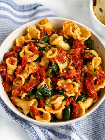 There is a white bowl filled with sun dried tomatoes, baby spinach, spices and herbs over cooked pasta.