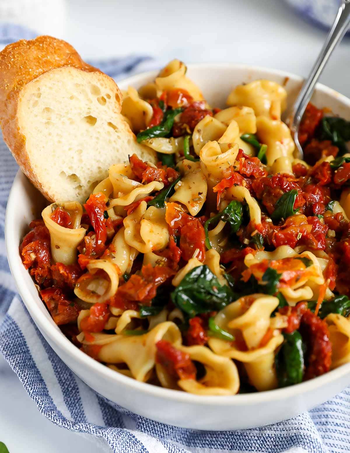 A fork placed inside a white bowl that is filled with pasta, tomatoes, and spinach. There is also a sliced of bread inside the bowl.