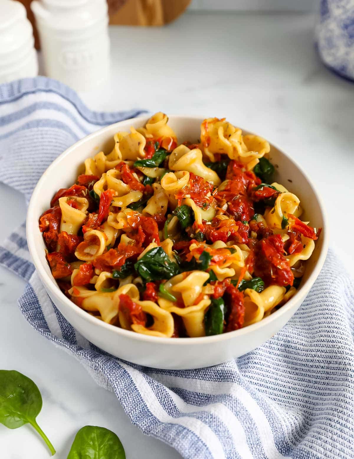 A white bowl filled with a large amount of cooked pasta with a tomato/spinach sauce on top.
