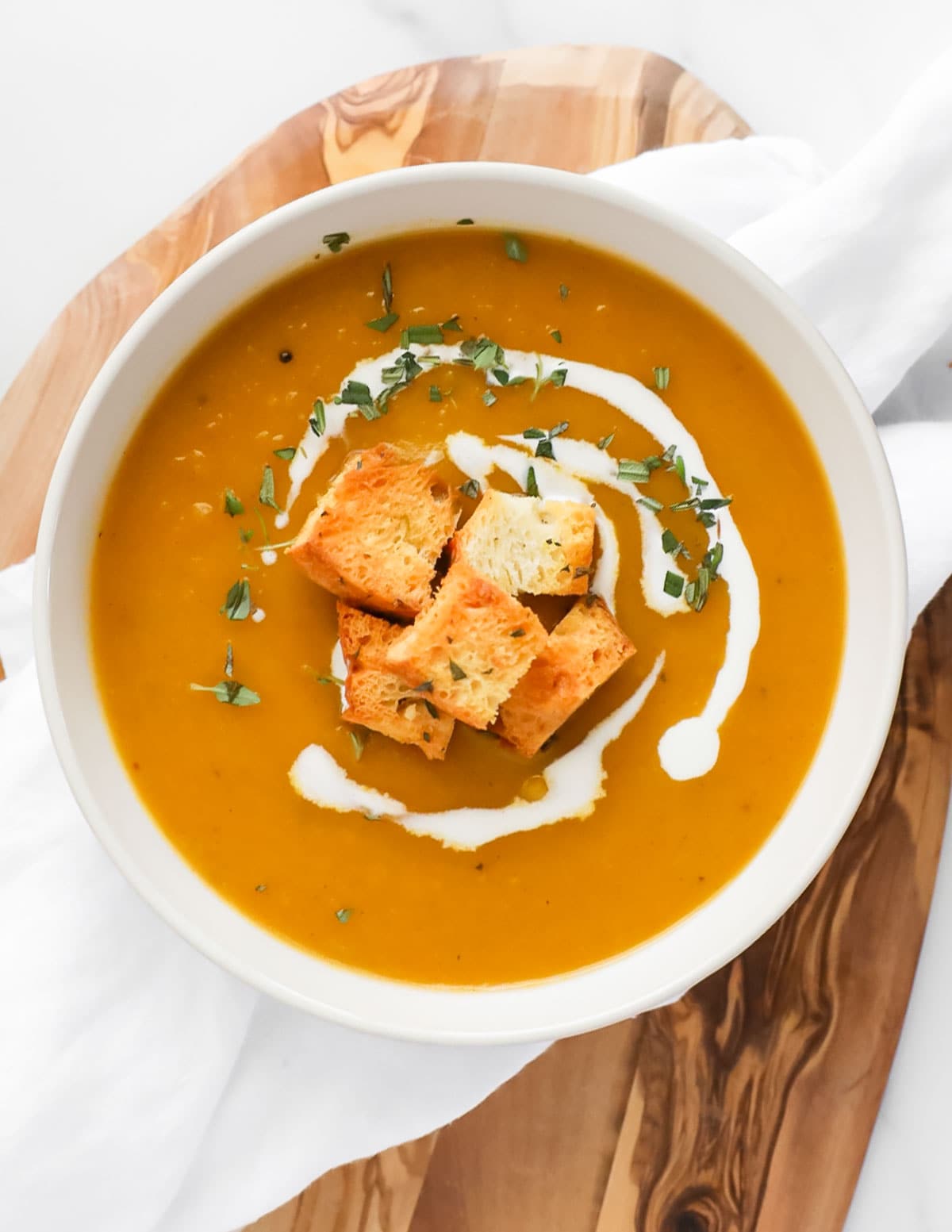 A white bowl filled with creamy orange soup with a drizzle of coconut milk on top along with five croutons, and chopped green herbs.