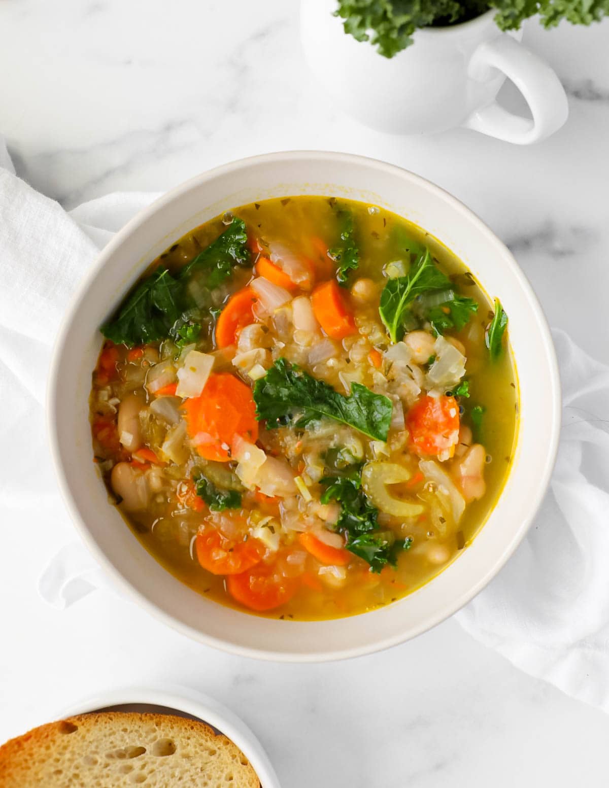 A white bowl filled with soup containing carrots, celery, beans, and kale. rrots, kale,
