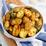 Golden yellow mini cooked baby potatoes in a white bowl with fresh herbs and seasoning on them.