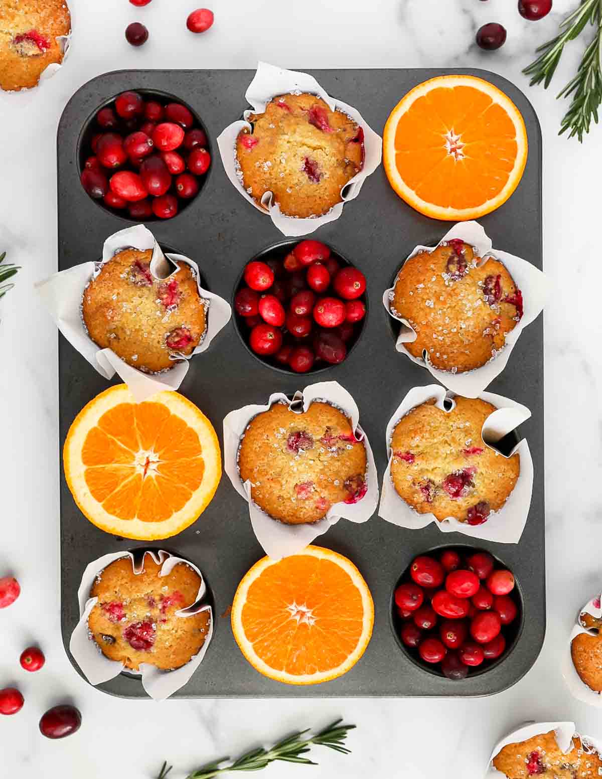 A muffin tin filled with baked muffins, cranberries, and sliced oranges.