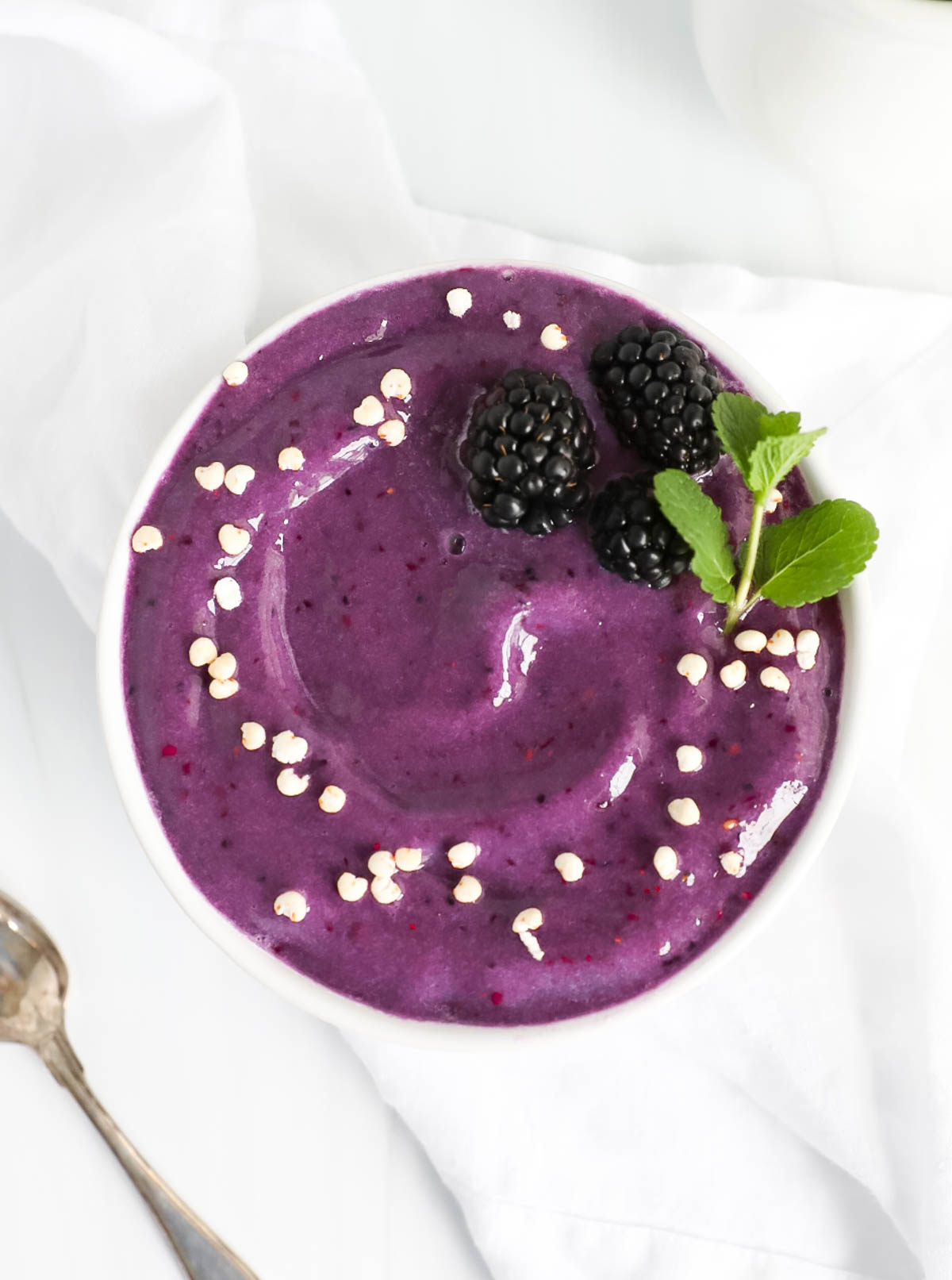 A purple smoothie in a white bowl garnished with three blackberries, fresh mint, and white puffed cereal.