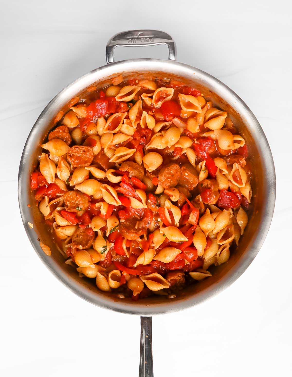 An overhead image of pasta in a large silver pan with sausages, tomatoes, peppers, and sauce.