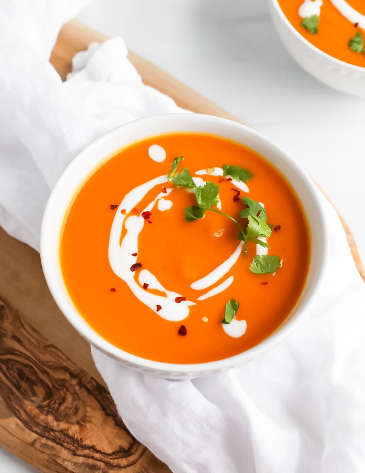 A white bowl filled with carrot soup that is topped with creamy coconut milk, fresh cilantro, and red pepper flakes.