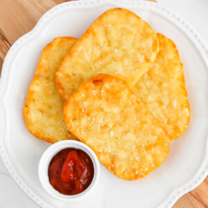 Four crispy hash browns on a plate with a small dish of ketchup.