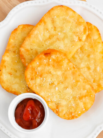 Four crispy hash browns on a plate with a small dish of ketchup.