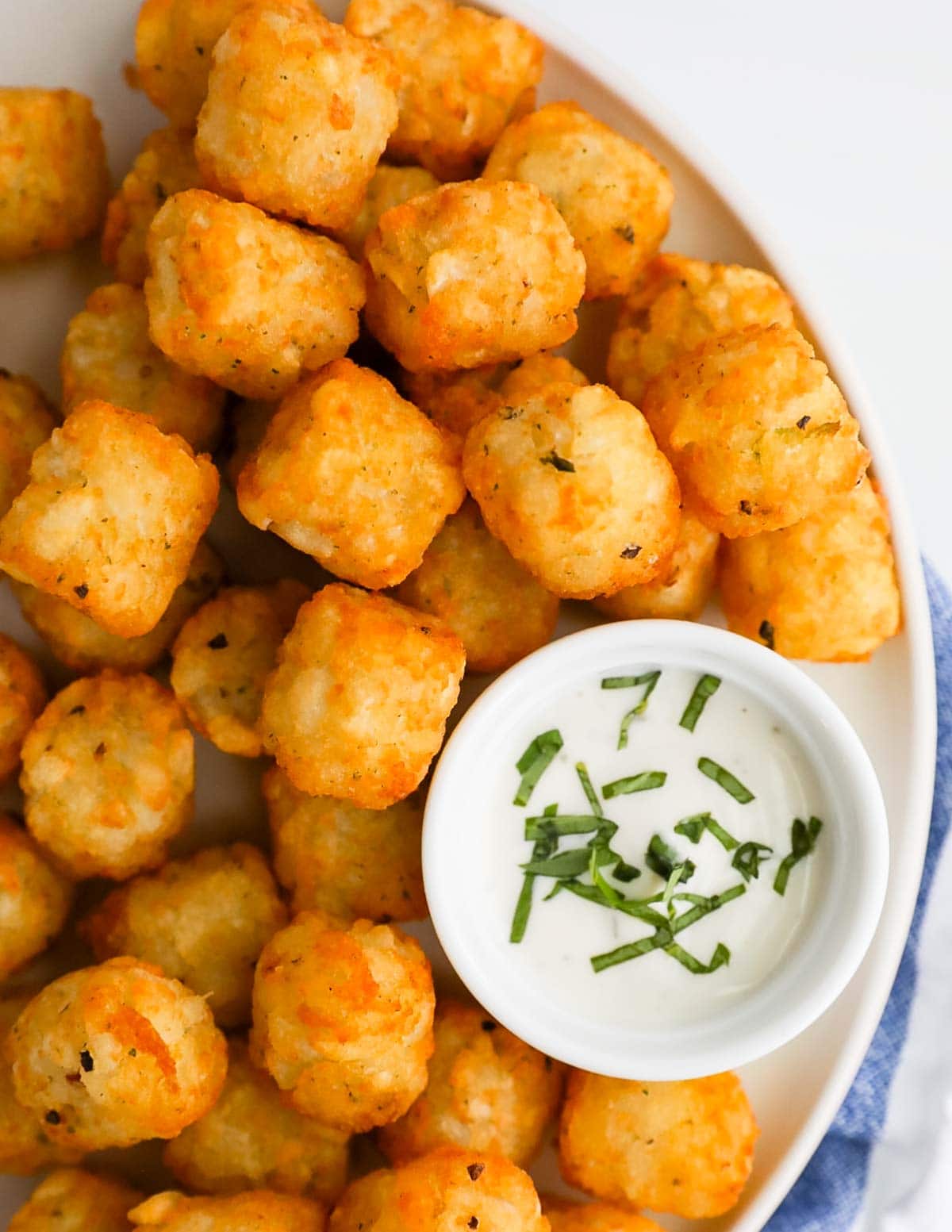 A close-up picture of tater tots on a white plate along with a white dish filled with ranch dressing and fresh basil.