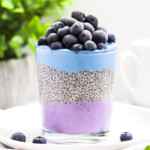 A glass filled with a layer of purple yogurt, chia pudding, blue yogurt, and blueberries on top.