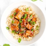 An overhead image of a white bowl filled with sauce-coated tofu over white rice. It is garnished with sesame seeds and green onion slices.