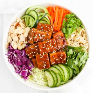 A white bowl filled with vegetables including: carrots, cucumbers, green onions, cilantro, green cabbage, red cabbage. It also has brown rice, almonds, and tofu with a brown sauce.