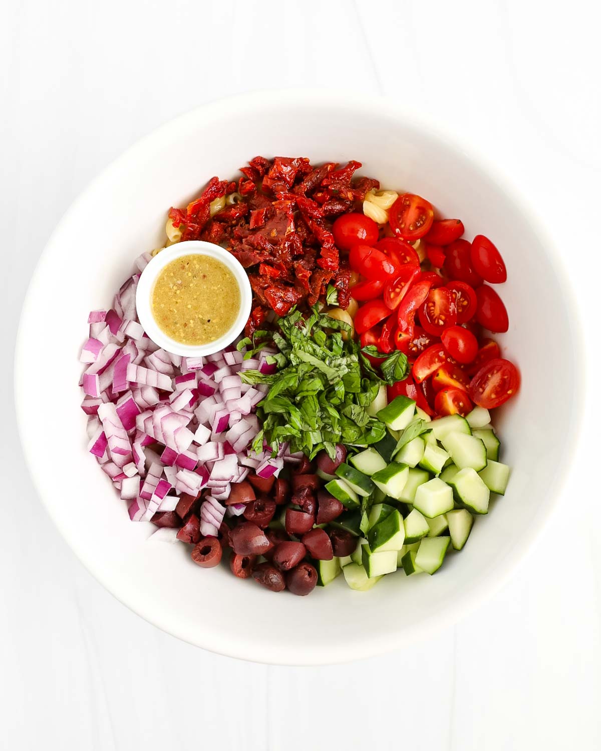A bowl with sections of ingredients including tomatoes, sun dried tomatoes, cucumbers, olives, red onion, basil, and a small white bowl of homemade salad dressing.