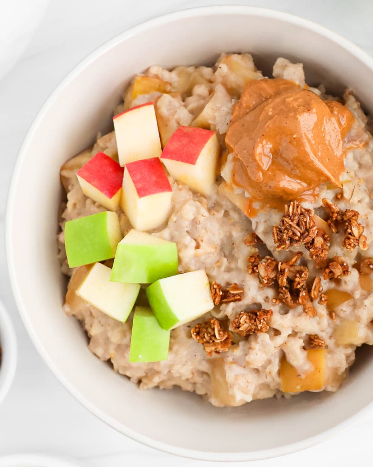 A overhead image of cooked apple oatmeal in a bowl covered with peanut butter, granola, and apples.