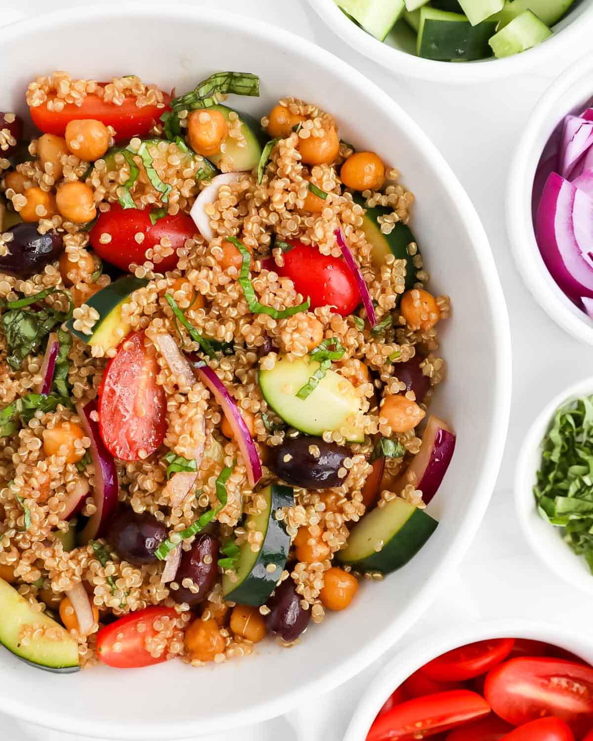 A white bowl filled with a mixture of quinoa, tomatoes, cucumber, olives, tomato, and onion.