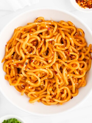 A white bowl of noodles that are covered in white sesame seeds and red pepper flakes.