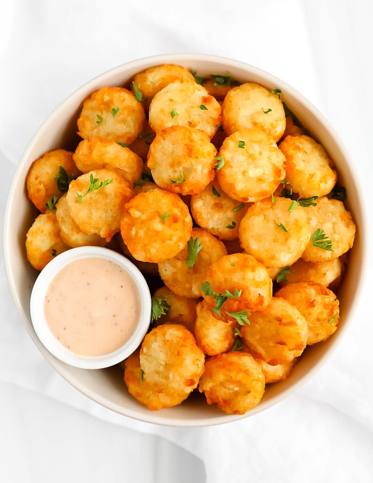 A white bowl filled with cooked tater tots and a small bowl of spicy ranch dressing. There is fresh parsley on top.