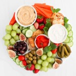A round white tray filled with a variety of fresh foods including: hummus, tzatziki, strawberries, grapes, figs, cucumbers, pickles, olives, peppers, crackers.