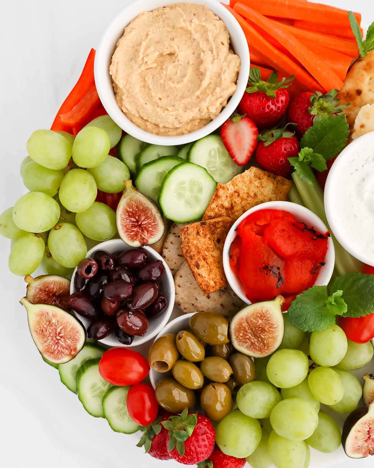 An overhead image of a spread of different foods including: hummus, peppers, olives, grapes, cucumbers, pita, strawberries, mint, and figs.