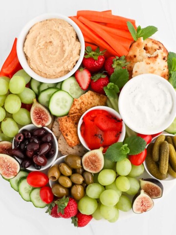 A round white tray filled with a variety of fresh foods including: hummus, tzatziki, strawberries, grapes, figs, cucumbers, pickles, olives, peppers, crackers.
