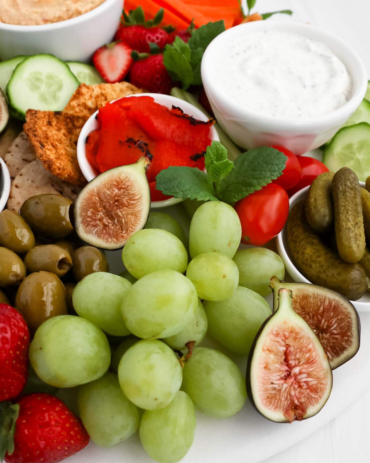A close up image of a charcuterie board with grapes, figs, olives, pickles, tomatoes, strawberries, tzatziki, crackers, and cucumbers.