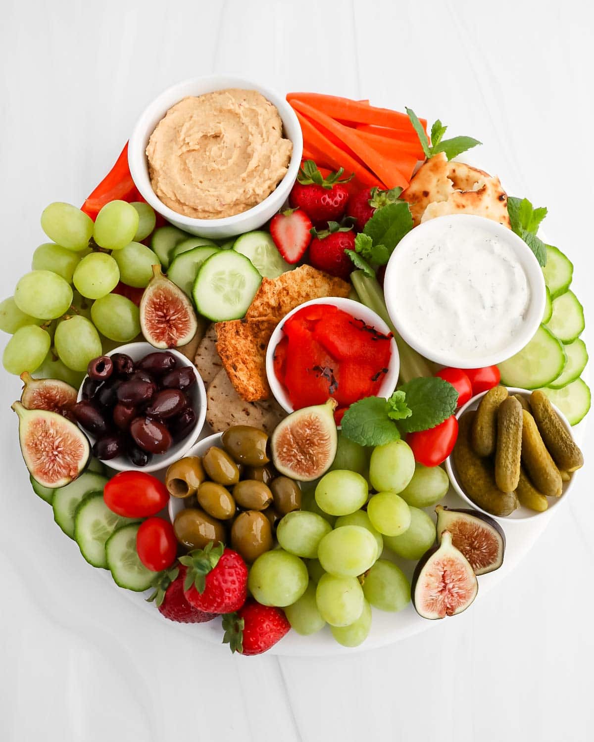 An overhead image of a white board covered in dips, peppers, cucumbers, tomatoes, pickles, olives, strawberries, grapes, and figs.