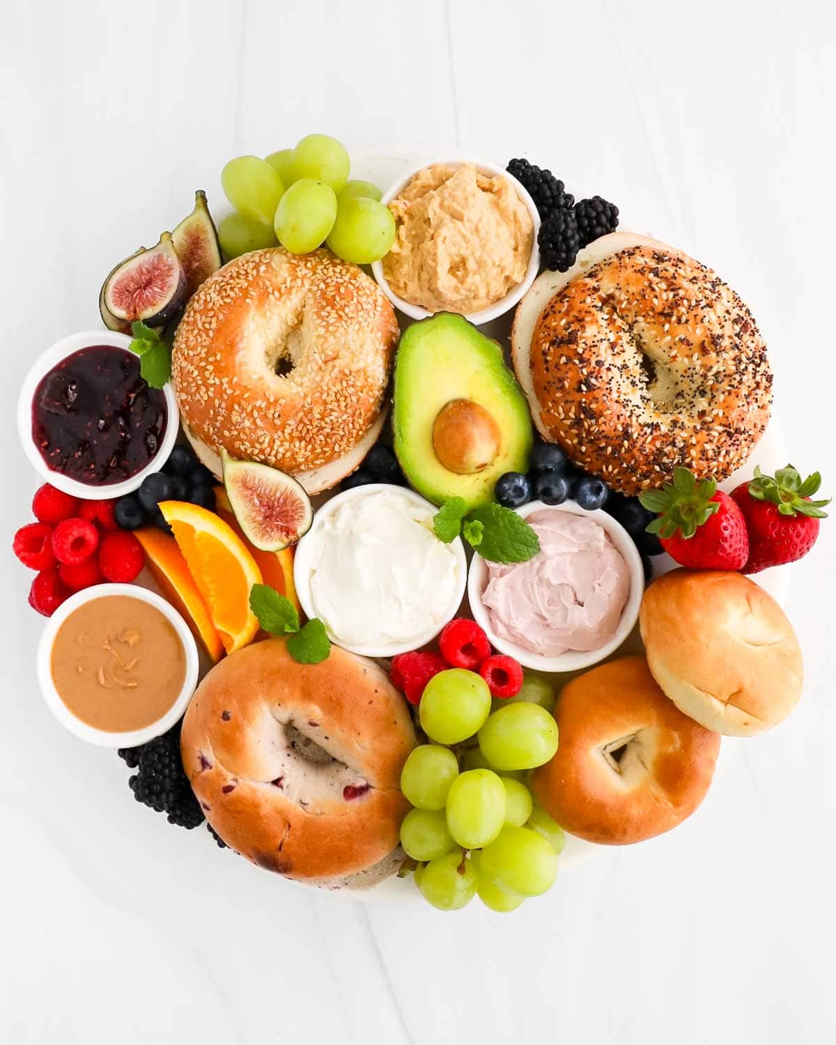 A board filled with different types of bagels, avocado, grapes, strawberries, raspberries, blueberries, oranges, blackberries, peanut butter, jelly, cream cheese, and fresh mint.