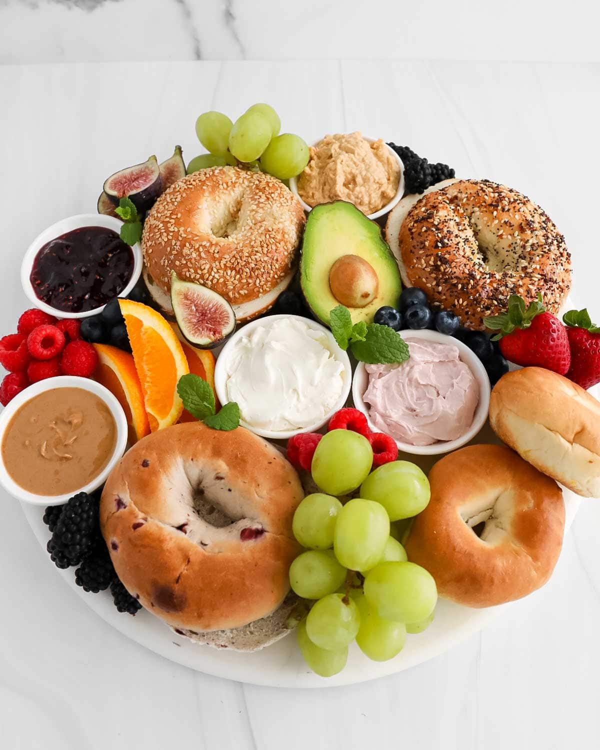 A round marble board filled with bagels, avocado, grapes, strawberries, raspberries, blueberries, blackberries, oranges, figs, mint, peanut butter, jelly, whipped cream, and creamy cheese.