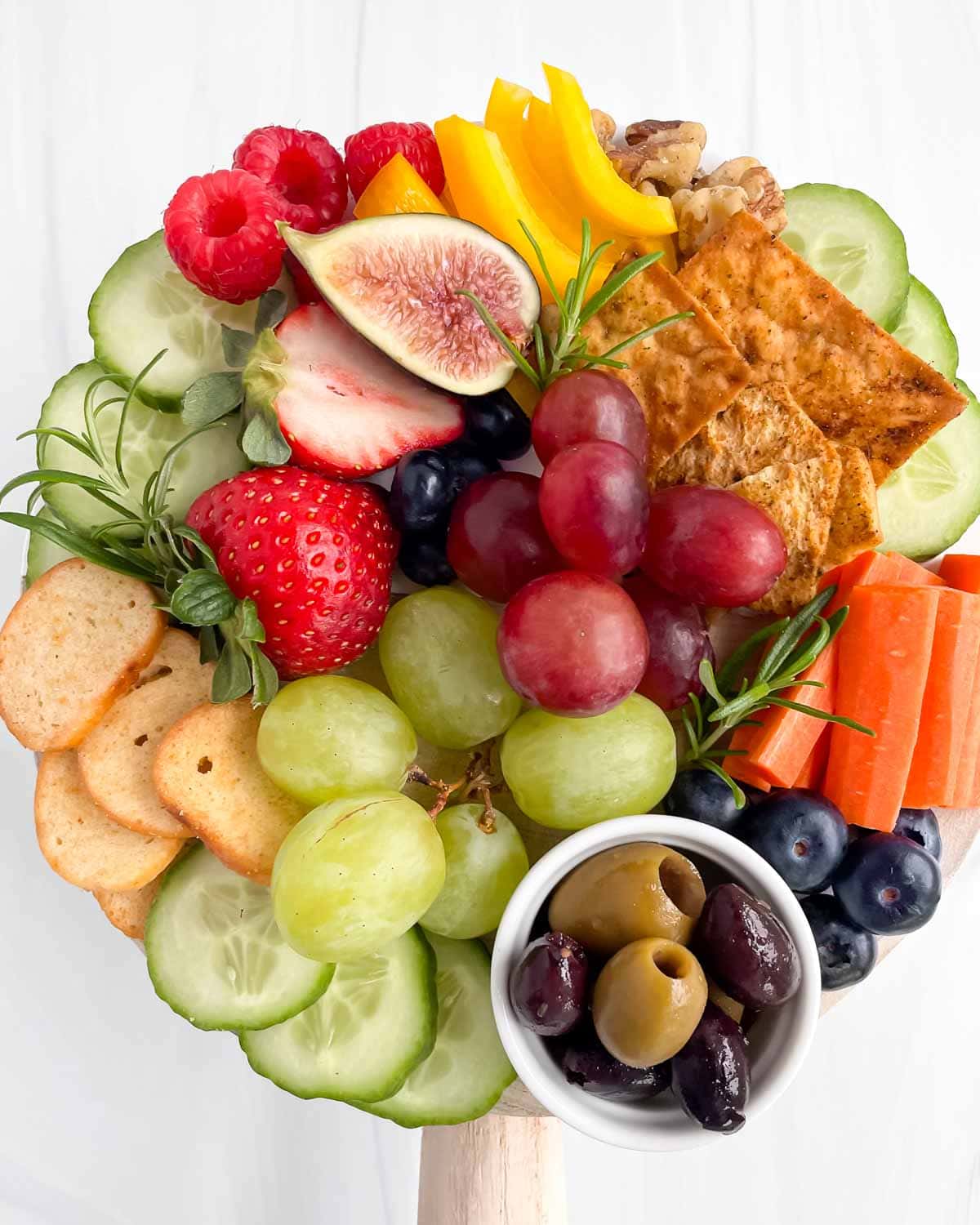 A small charcuterie board filled with olives, blueberries, strawberries, raspberries, grapes, cucumbers, carrots, peppers, crackers, and fresh rosemary.