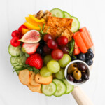 An overhead image of a wooden board covered in grapes, cucumbers, blueberries, strawberries, raspberries, olives, figs, walnuts, peppers, and crackers.